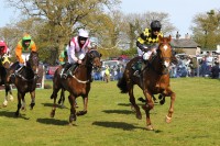 Bild von einem Pferderennen mit Jockeys in bunten Rennfarben reitend auf Pferden vor Zuschauern